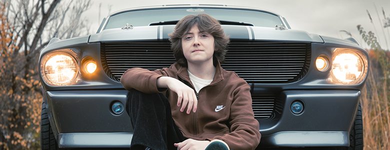 A high school senior with medium-length hair poses in front of a vintage car, its glowing headlights providing a cinematic touch to their senior pictures. Clad in a brown jacket and dark pants, they are framed by autumn foliage and a cloudy sky.