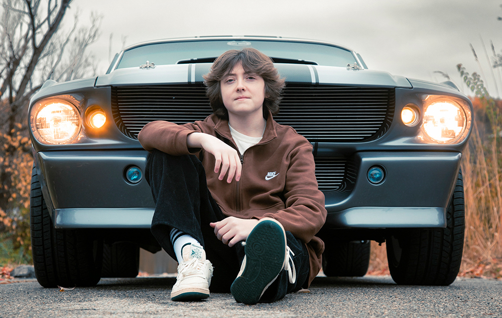 A person with shoulder-length hair is seated on the ground, posing for senior pictures in front of a classic car with its headlights aglow. Dressed in a brown jacket, black pants, and white sneakers, they sit outdoors under overcast skies, surrounded by bare trees.
