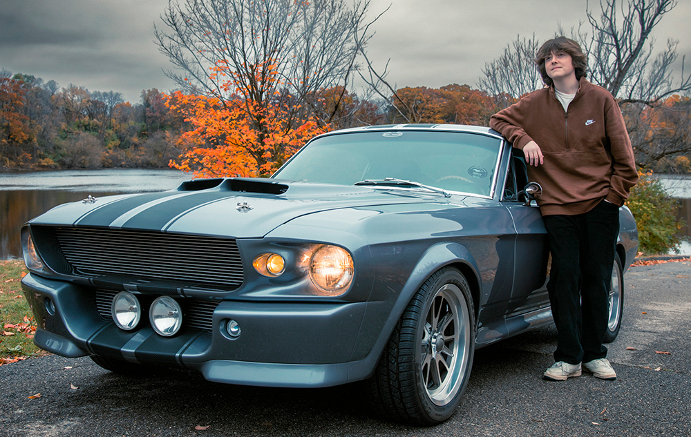 A person in a brown jacket and black pants poses for senior pictures, leaning casually against a vintage grey car with racing stripes. The autumn backdrop features colorful trees and a serene lake, adding depth to this timeless scene.