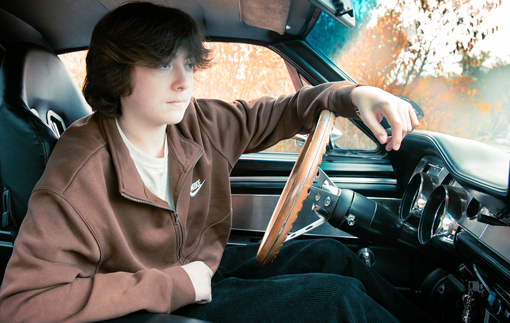A high school senior with brown hair sits in a car, posing for senior pictures with cars as they rest an arm on the steering wheel. They wear a brown jacket and a white shirt, perfectly complementing the dark interior. Through the window, trees blur by, framing this timeless moment.