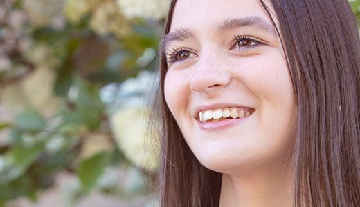 A high school senior girl smiles joyfully, standing among blooming white hydrangeas. Her light pink dress contrasts with the lush greenery and soft white flowers, capturing a beautiful moment for her senior pictures.