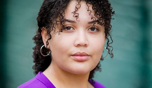 A child actor headshot of a girl with curly hair, wearing a purple top and hoop earrings, against a green background. She has a subtle, serene expression.