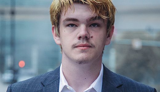 A high school senior boy with blonde hair and a light mustache stands in an urban setting, perfect for foggy senior pictures. He wears a white shirt and a grey plaid blazer. The background is blurred, featuring buildings, a traffic light, and light fixtures on a misty day.