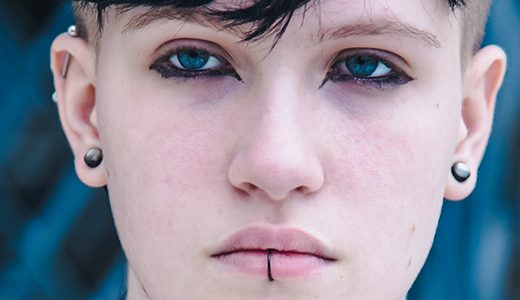 Close-up of a goth high school senior with short, dark hair and blue eyes, wearing black eyeliner and having piercings in their ears and below their lower lip. The background is blurry with a blue shade, making the person's face the focal point.
