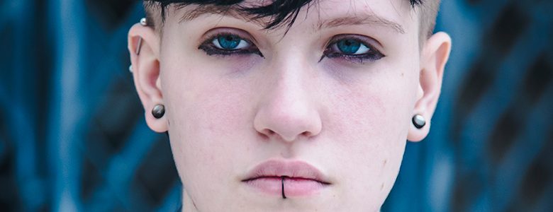 Close-up of a goth high school senior with short, dark hair and blue eyes, wearing black eyeliner and having piercings in their ears and below their lower lip. The background is blurry with a blue shade, making the person's face the focal point.