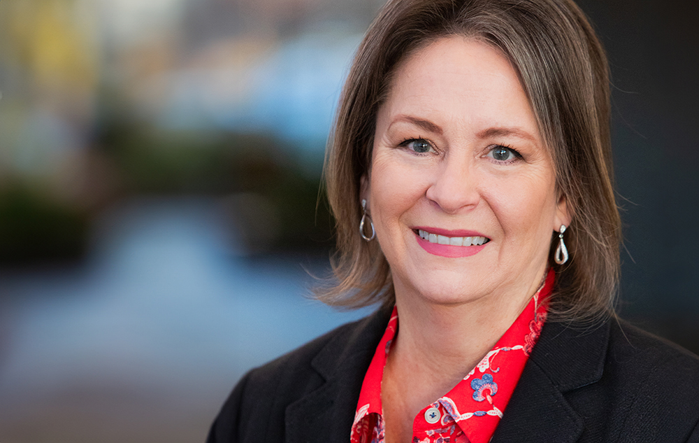 A professional woman with a friendly smile, wearing a black jacket and red scarf, poses in an office with a blurred background for a professional LinkedIn headshot in Detroit, MI.