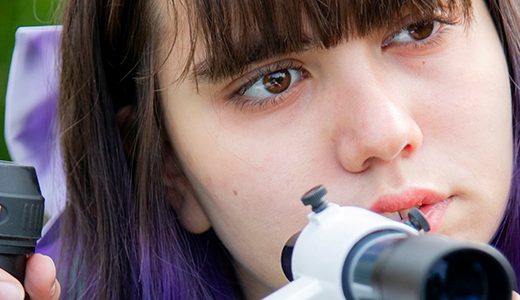 A high school senior girl with purple hair looking through a telescope's viewfinder, focusing intently on aligning it, with green trees blurred in the background.