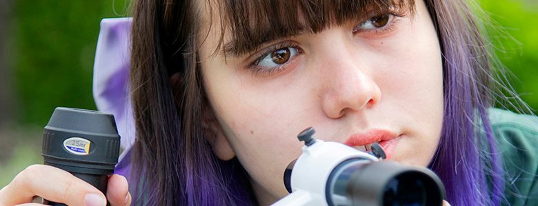 A high school senior girl with purple hair looking through a telescope's viewfinder, focusing intently on aligning it, with green trees blurred in the background.