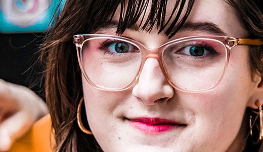 Close-up of a smiling young woman wearing glasses and dangling earrings, with colorful skateboard artwork visible in the background.