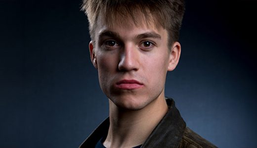 A young man with short, light brown hair stares directly at the camera against a dark background. He is wearing a dark jacket and appears serious, with a neutral expression on his face. The low lighting creates a dramatic effect, ideal for Detroit Actor Headshots.