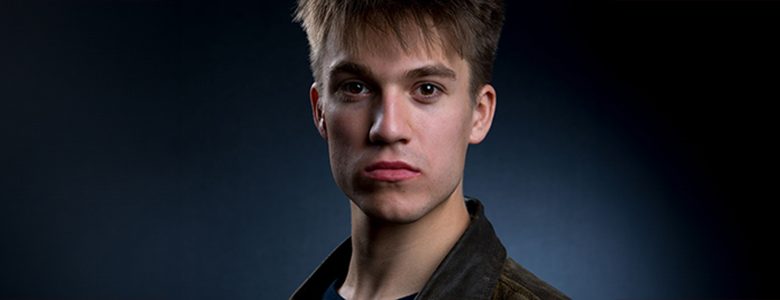 A young man with short, light brown hair stares directly at the camera against a dark background. He is wearing a dark jacket and appears serious, with a neutral expression on his face. The low lighting creates a dramatic effect, ideal for Detroit Actor Headshots.