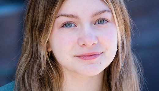 A child actress with long, straight hair and a gentle expression is looking at the camera. She is wearing a light-colored shirt and a grey cardigan. The photo, ideal for child acting headshots, has a watermark from Paul Manoian Photography at the bottom left corner.