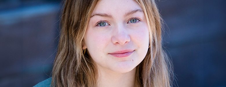 A child actress with long, straight hair and a gentle expression is looking at the camera. She is wearing a light-colored shirt and a grey cardigan. The photo, ideal for child acting headshots, has a watermark from Paul Manoian Photography at the bottom left corner.