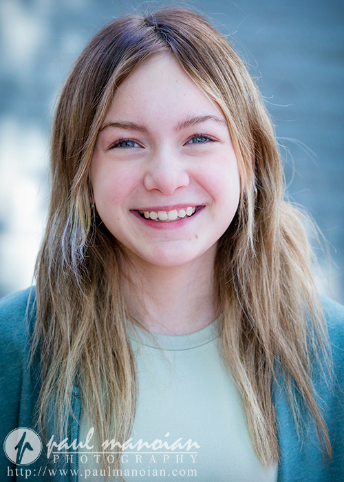 A child actress with long, light brown hair smiles at the camera. She is wearing a light-colored shirt and a darker-colored cardigan. The background is blurred and bright, directing focus on the individual. This captivating shot is perfect for child acting headshots, with a logo for Paul Manoian Photography at the bottom.
