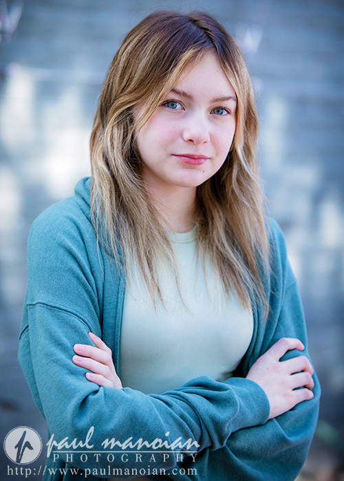 A child actor with long, light brown hair stands outdoors with their arms crossed, wearing a light green shirt and a blue cardigan. They have a neutral expression. The background is blurred, capturing the essence of natural light lifestyle child acting headshots, and the image bears the watermark of Paul Manoian Photography at the bottom.