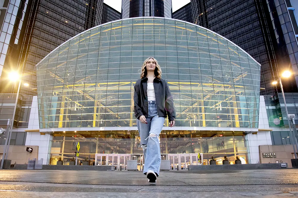 Detroit Senior Pictures A high school senior girl wearing a black jacket and ripped jeans walks on a plaza in front of the Renaissance Center for her Detroit senior pictures session. The scene is lit by streetlights, enhancing the modern architectural features.