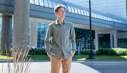 A high school senior guy wearing a light green shirt and beige pants stands with hands in pockets, smiling and looking to the side. They are outdoors in a modern urban area of downtown Detroit, with a glass bridge, buildings, trees, and green plants in the background—perfect for senior pictures.