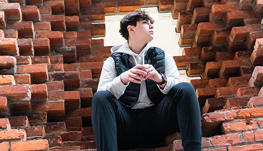 A high school senior boy with short dark hair, wearing a black vest over a grey hoodie and black pants, is seated within a large, irregular hole in a brick wall. They appear to be looking thoughtfully into the distance for their downtown Detroit senior pictures session with Paul Manoian Photography.