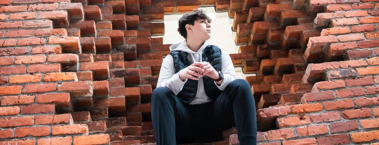 A high school senior boy with short dark hair, wearing a black vest over a grey hoodie and black pants, is seated within a large, irregular hole in a brick wall. They appear to be looking thoughtfully into the distance for their downtown Detroit senior pictures session with Paul Manoian Photography.