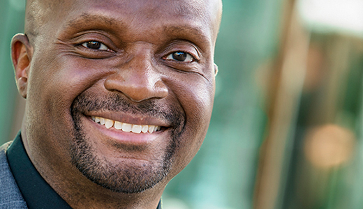 A man with a bald head and goatee is smiling warmly at the camera in this professional LinkedIn headshot. He is wearing a dark jacket and a black shirt. The background is blurred and features greens and browns.