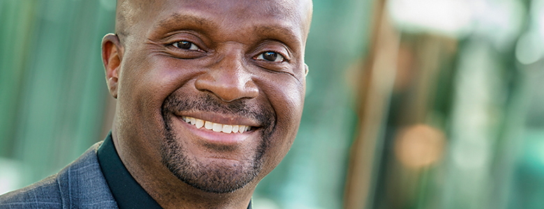 A man with a bald head and goatee is smiling warmly at the camera in this professional LinkedIn headshot. He is wearing a dark jacket and a black shirt. The background is blurred and features greens and browns.