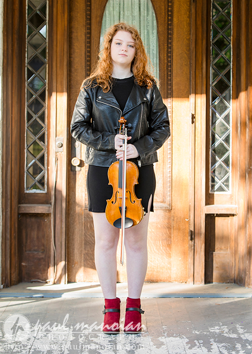 North Farmington Senior Pictures A high school senior girl with long, curly red hair stands in front of a wooden door, holding a violin. She is dressed in a black dress, black leather jacket, and red boots for her North Farmington senior pictures session with Paul Manoian Photography.