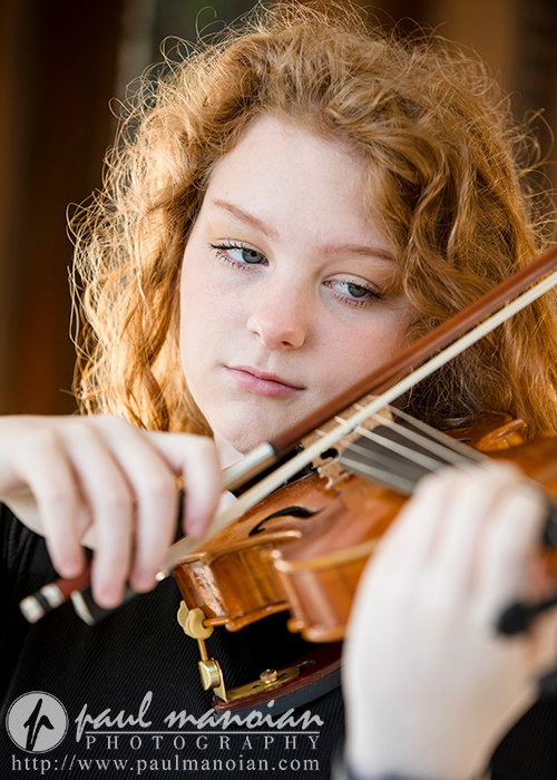 North Farmington Senior Pictures A high school senior girl with curly red hair plays a violin, focusing intently on the instrument. They wear a dark top, and the background is softly blurred for her North Farmington Senior Pictures session with Paul Manoian Photography.