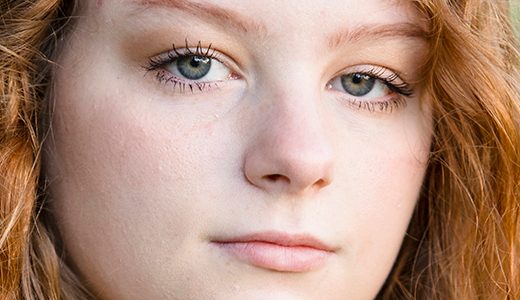 A close-up of a high school senior girl with light skin and long, wavy red hair for her North Farmington senior pictures session with Paul Manoian Photography. They have green eyes and a neutral facial expression. The background is blurred with green tones, suggesting an outdoor setting.