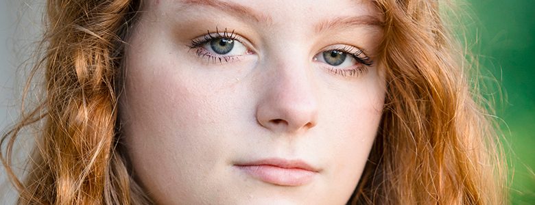 A close-up of a high school senior girl with light skin and long, wavy red hair for her North Farmington senior pictures session with Paul Manoian Photography. They have green eyes and a neutral facial expression. The background is blurred with green tones, suggesting an outdoor setting.