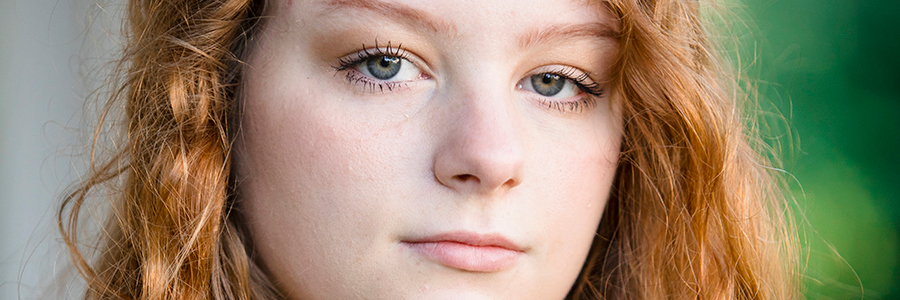 A close-up of a high school senior girl with light skin and long, wavy red hair for her North Farmington senior pictures session with Paul Manoian Photography. They have green eyes and a neutral facial expression. The background is blurred with green tones, suggesting an outdoor setting.