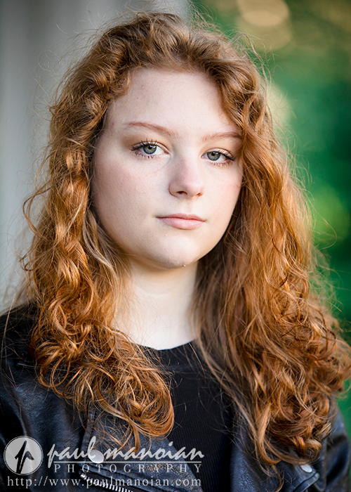 North Farmington Senior Pictures A high school senior girl with long, curly red hair and fair skin gazes at the camera with a slight, neutral expression for her North Farmington senior pictures session with Paul Manoian Photography. They are wearing a black top, and the background is softly blurred greenery.