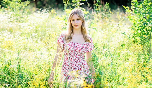 A high school senior girl in a white and red floral dress stands amidst tall grass and wildflowers in a sunny meadow, capturing the essence of Ann Arbor Senior Pictures. With her long blonde hair flowing and sunlight filtering through lush foliage, she gazes directly at the camera.
