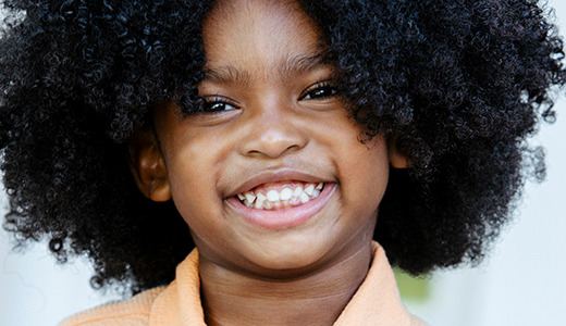A young child with curly dark hair, smiling brightly, and wearing an orange shirt. The background is slightly blurred, indicating an outdoor setting with some buildings or structures behind. This delightful image captures the charm often seen in kids modeling headshots.