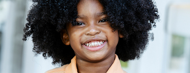 A young child with curly dark hair, smiling brightly, and wearing an orange shirt. The background is slightly blurred, indicating an outdoor setting with some buildings or structures behind. This delightful image captures the charm often seen in kids modeling headshots.