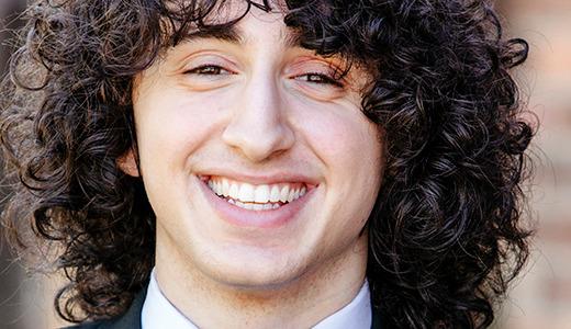 A high school senior with curly dark hair and a bright smile, dressed in a formal suit and tie, is posing outdoors for their Macomb senior pictures against the backdrop of a blurred brick wall.