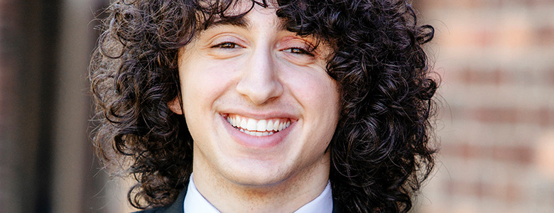 A high school senior with curly dark hair and a bright smile, dressed in a formal suit and tie, is posing outdoors for their Macomb senior pictures against the backdrop of a blurred brick wall.