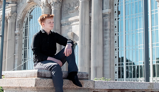 A high school senior in a black outfit sits on a stone railing in front of the historic Michigan Central Station building with large arched windows and columns, taking their U of D Jesuit senior pictures. The sky is clear and blue, highlighting the intricate architectural details behind them.