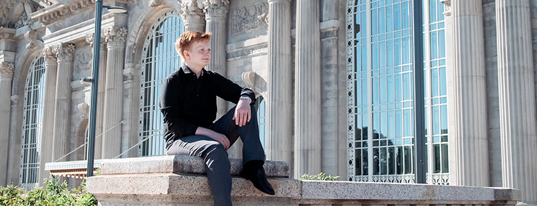 A high school senior in a black outfit sits on a stone railing in front of the historic Michigan Central Station building with large arched windows and columns, taking their U of D Jesuit senior pictures. The sky is clear and blue, highlighting the intricate architectural details behind them.