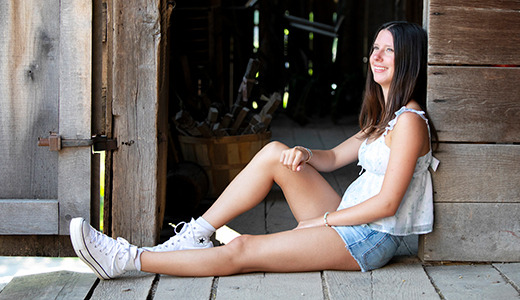 A high school senior with long hair sits on the floor in a doorway of a rustic wooden building, perfect for senior portraits. She wears a light sleeveless top, denim shorts, and white sneakers, and she is smiling while looking off to the side. The background is dimly lit and features old wooden structures.