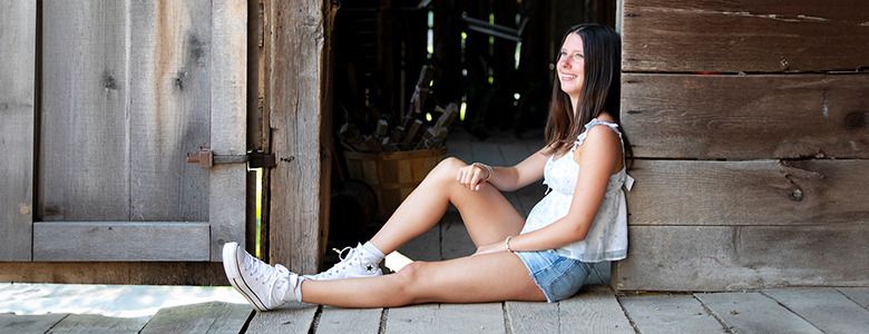 A high school senior with long hair sits on the floor in a doorway of a rustic wooden building, perfect for senior portraits. She wears a light sleeveless top, denim shorts, and white sneakers, and she is smiling while looking off to the side. The background is dimly lit and features old wooden structures.