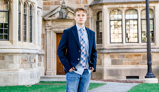A high school senior stands confidently outside a large stone building with arched windows and a wooden door, perfect for Ann Arbor senior portraits. They are wearing a dark blazer over a light shirt with a tie, blue jeans, and white sneakers, with their hands in their pockets. The grassy area and pathway frame the scene perfectly.