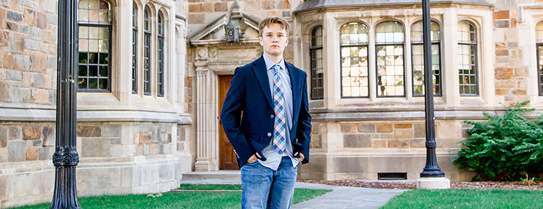 A high school senior stands confidently outside a large stone building with arched windows and a wooden door, perfect for Ann Arbor senior portraits. They are wearing a dark blazer over a light shirt with a tie, blue jeans, and white sneakers, with their hands in their pockets. The grassy area and pathway frame the scene perfectly.