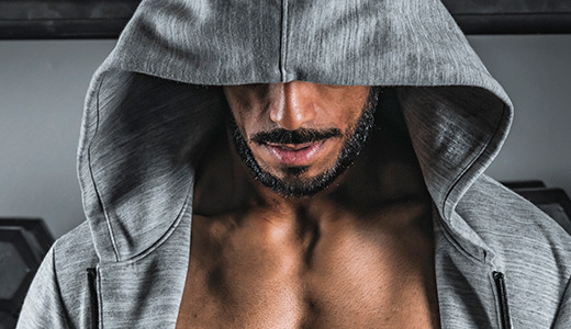 A person wearing a gray hoodie with the hood covering their eyes. They have a beard and mustache, and their muscular chest, honed by Crossfit, is visible. Behind them are hexagonal dumbbells arranged on a rack. The background is a plain wall.