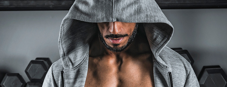 A person wearing a gray hoodie with the hood covering their eyes. They have a beard and mustache, and their muscular chest, honed by Crossfit, is visible. Behind them are hexagonal dumbbells arranged on a rack. The background is a plain wall.