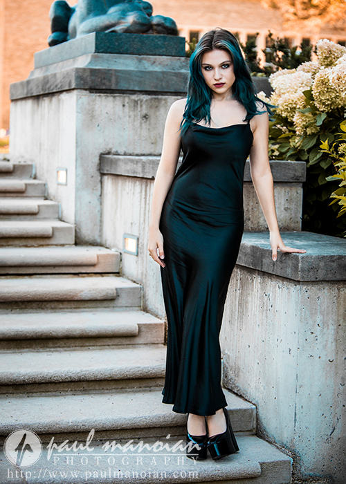 A high school senior in a sleek black dress and heels poses on an outdoor stone staircase. She leans against the railing, with one hand resting on it. Behind her are flowering bushes and a lion statue. The photographer's watermark, "Paul Manoian Photography," is visible at the bottom.