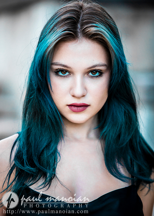 A high school senior with long blue hair and piercing eyes stares directly into the camera. They are wearing a black top and have a serious expression for a dark and moody senior pictures session. The image has a soft, blurred background. Photography branding and website information are visible at the bottom.