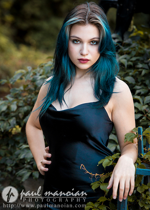 A high school senior with teal-colored hair and wearing a sleeveless black dress stands confidently in front of lush green foliage. She gazes directly at the camera with a serious expression, one hand resting on a blue railing. The image features the logo of Paul Manoian Photography.