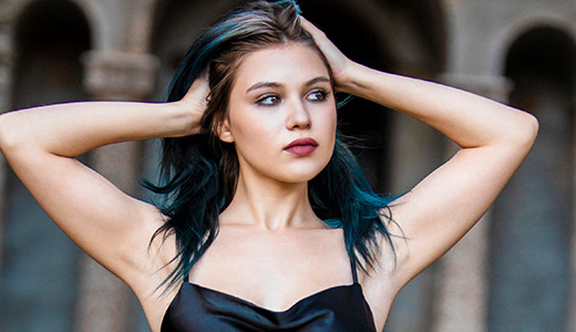 A high school senior with blue-tinted hair stands outdoors with her hands resting on her head. She is wearing a black strappy top and gazes off into the distance, evoking the feel of dark and moody senior pictures, with an architectural structure and blurred greenery in the background.