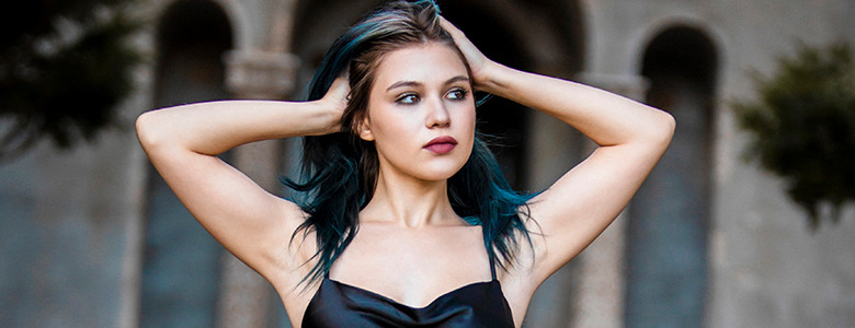 A high school senior with blue-tinted hair stands outdoors with her hands resting on her head. She is wearing a black strappy top and gazes off into the distance, evoking the feel of dark and moody senior pictures, with an architectural structure and blurred greenery in the background.