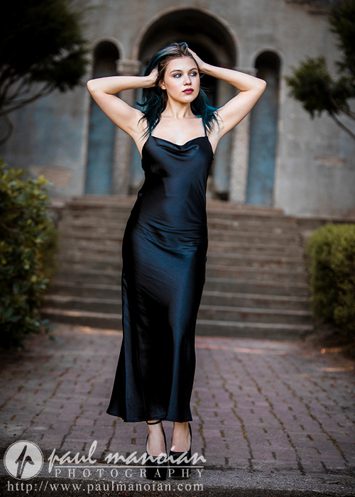A high school senior with dark hair in a long black dress stands on a brick pathway with her hands behind her head. She is posing in front of a stone building with steps leading up to it, and there are bushes on either side of the path. The image is watermarked with "Paul Manoian Photography.